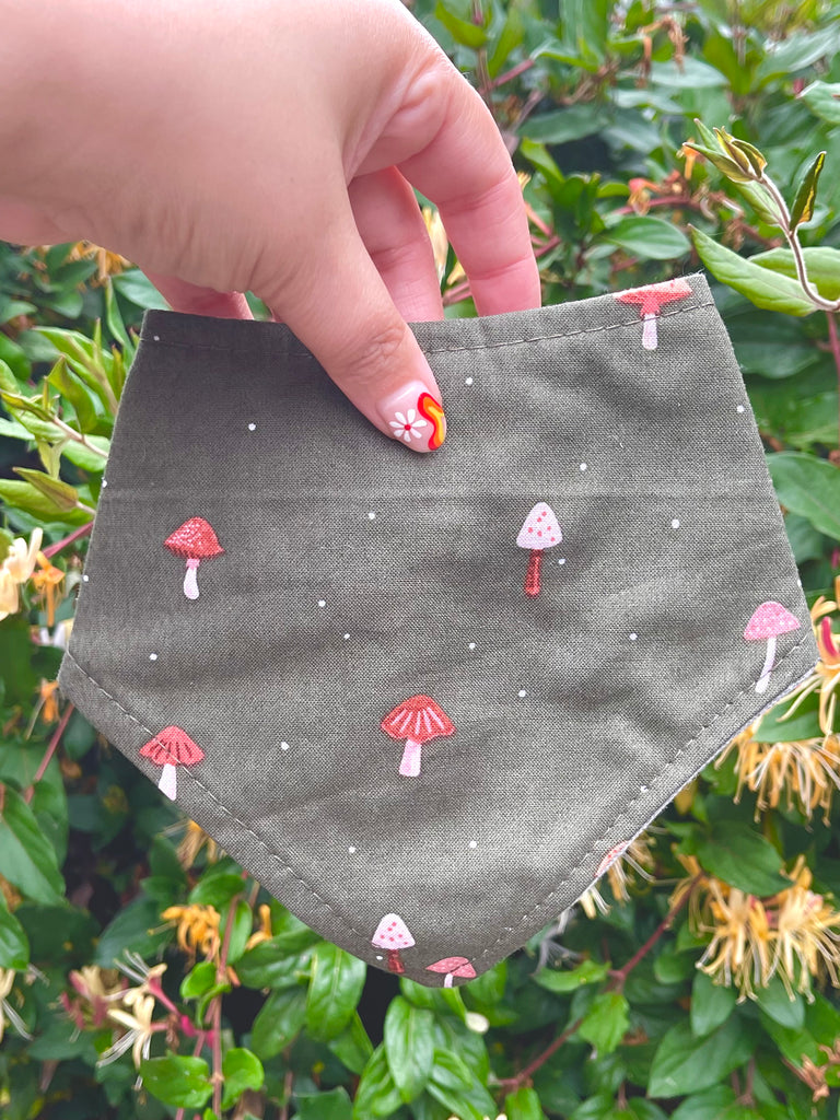 Olive green mushroom print dog bandana held up by hand in front of green leafy garden, showing full design. Perfect for fall or thanksgiving.