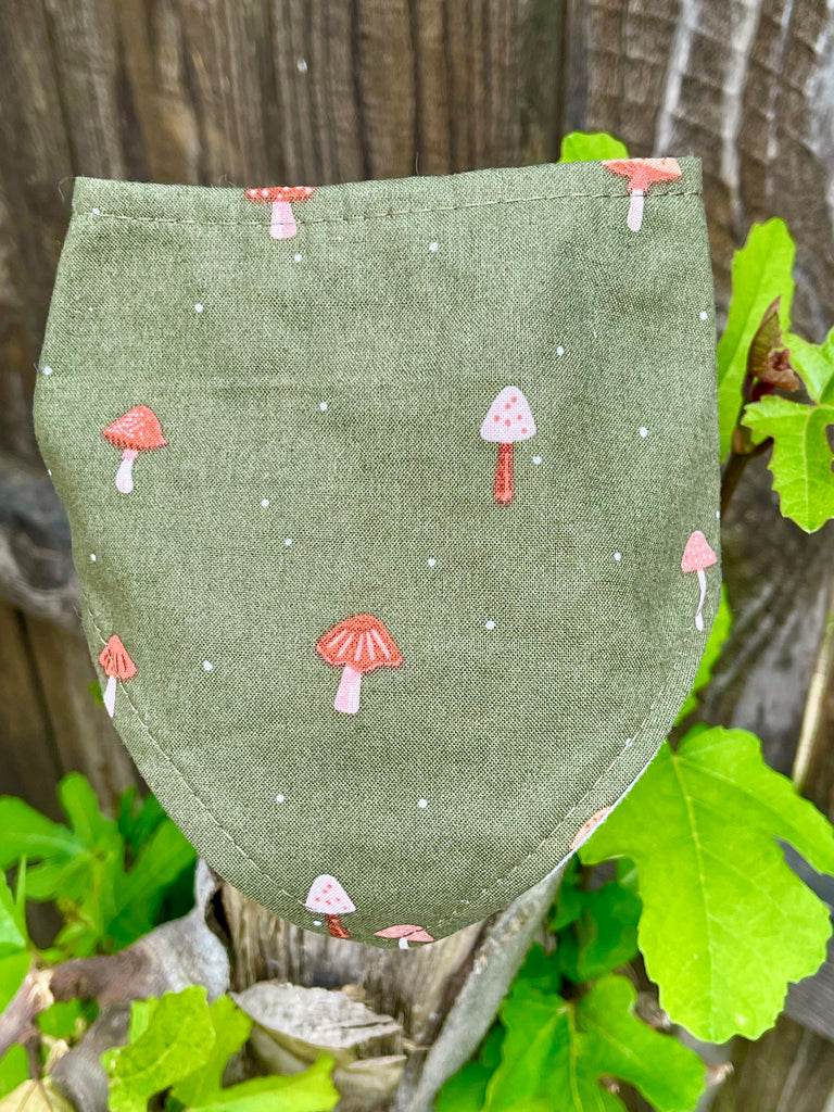 Close-up of olive green mushroom pet bandana displayed on wooden fence post, highlighting red toadstool pattern. Handmade in Los Angeles, California.
