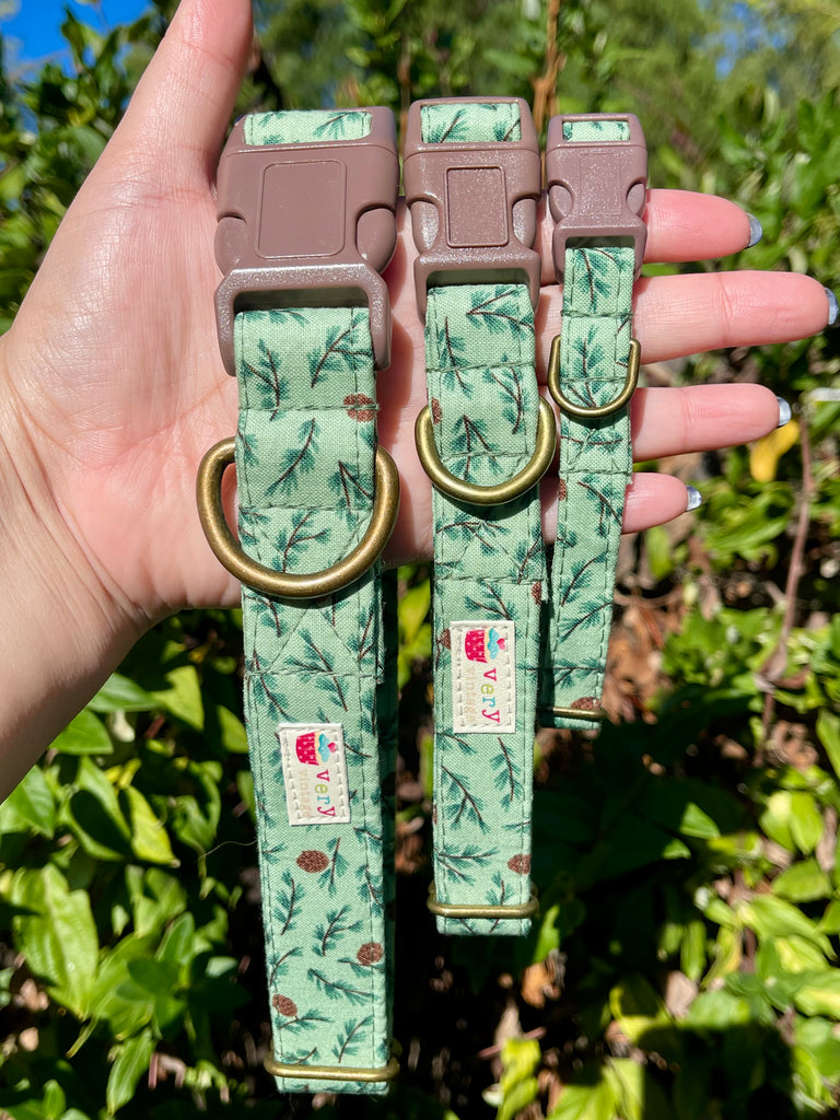 Hand holding three green Forest Pines collars in front of greenery – handmade holiday dog collars with pine branch print and brass hardware