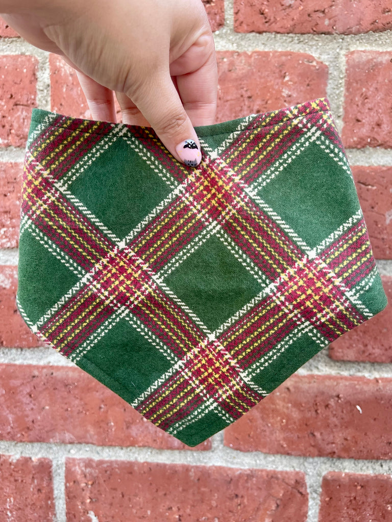 Hand holding a holiday dog bandana with a green, red, and yellow plaid pattern. The bandana is displayed against a brick wall backdrop.