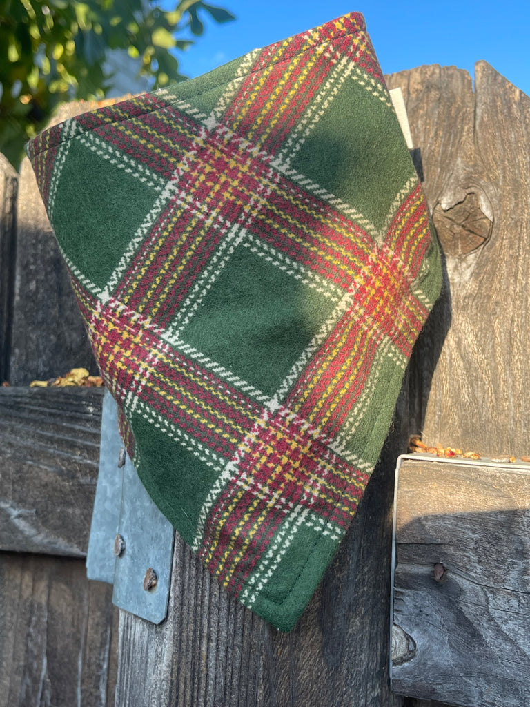Classic flannel dog bandana resting on a rustic wood fence in warm light. Plaid fabric shows green, cranberry, and gold tones.