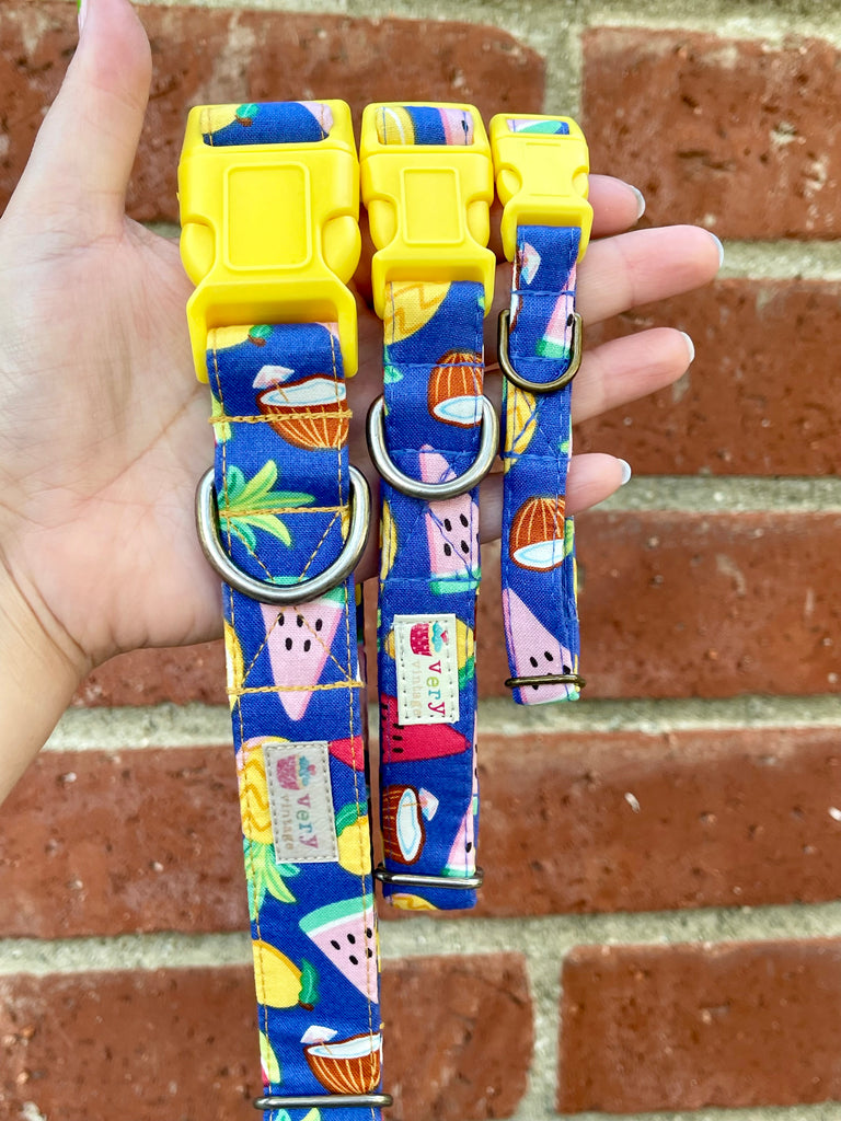 Three handmade dog collars with tropical fruit designs shown against a vintage red brick wall. Each collar has a yellow buckle and features fruits like watermelon and coconut, perfect for summer styling.