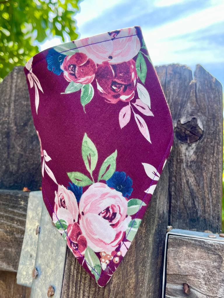 Dog bandana with blush and burgundy roses on plum background, hanging on rustic wood fence with greenery and sky in background