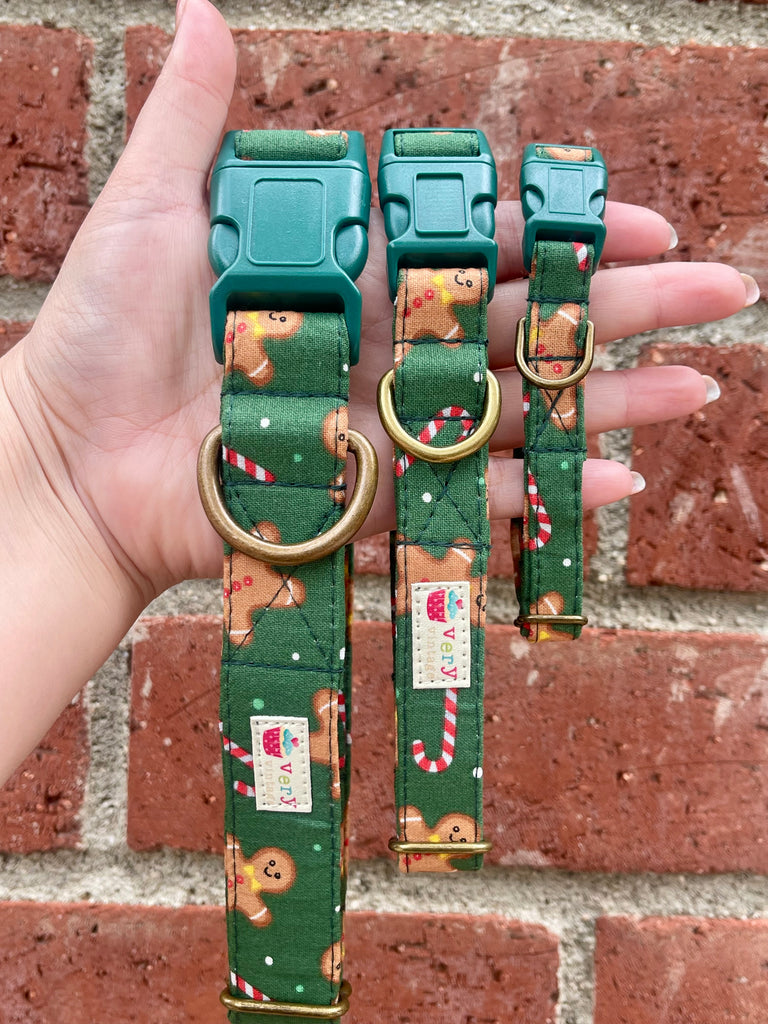 Hand holding three green holiday dog collars with gingerbread cookie and candy cane pattern against red brick wall background