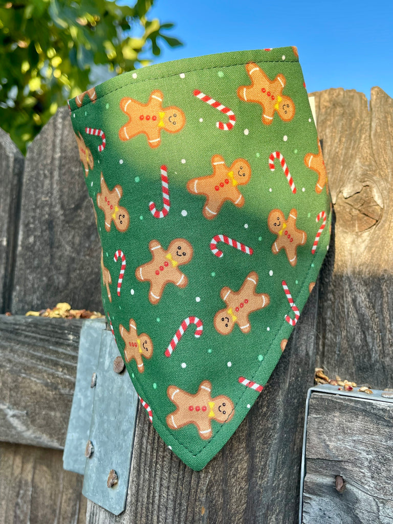 Gingerbread bandana draped over a rustic wooden fence, lit with warm morning sunlight. Features gingerbread men, candy canes, and snowy accents on green fabric.