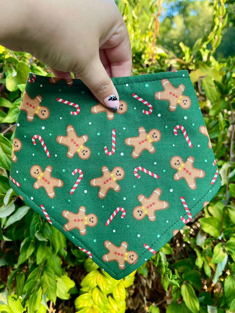 Close-up of the Gingerbread bandana held against lush greenery. Design shows smiling cookies, candy canes, and snow-like dots on a dark green background.