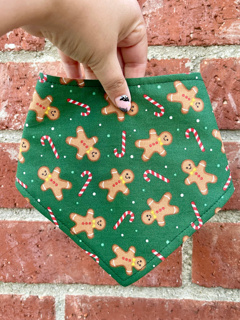 Hand holding a green dog bandana in front of a red brick wall. The bandana features gingerbread men and candy canes with white dot “snowflakes.”
