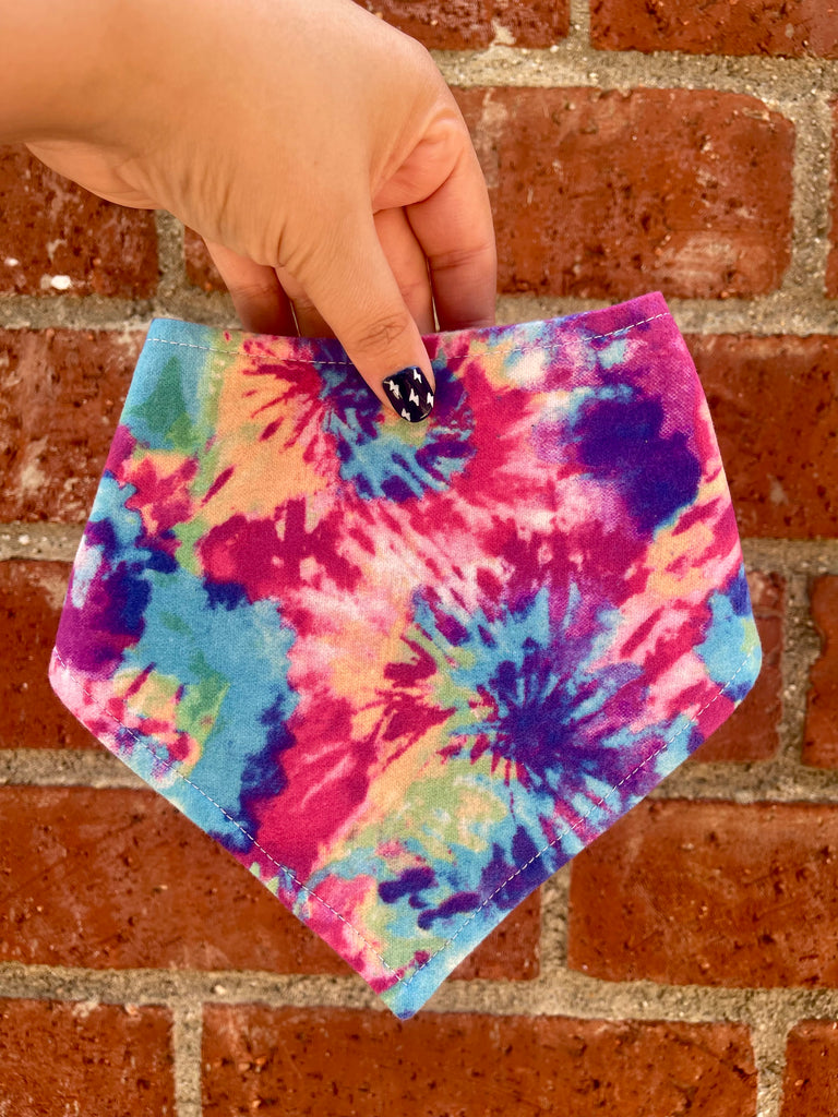 Hand holding tie-dye dog bandana with hot pink, blue, and purple swirls in front of a red brick wall