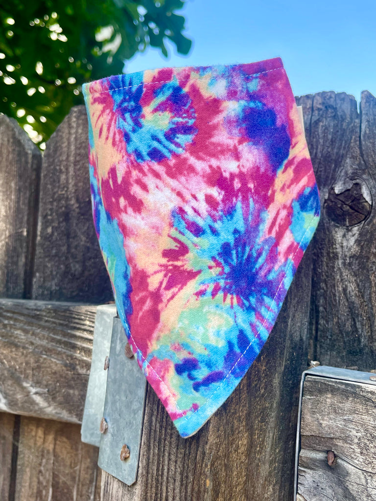 Vibrant tie-dye bandana displayed on a wood fence with tree leaves and blue sky behind it.
