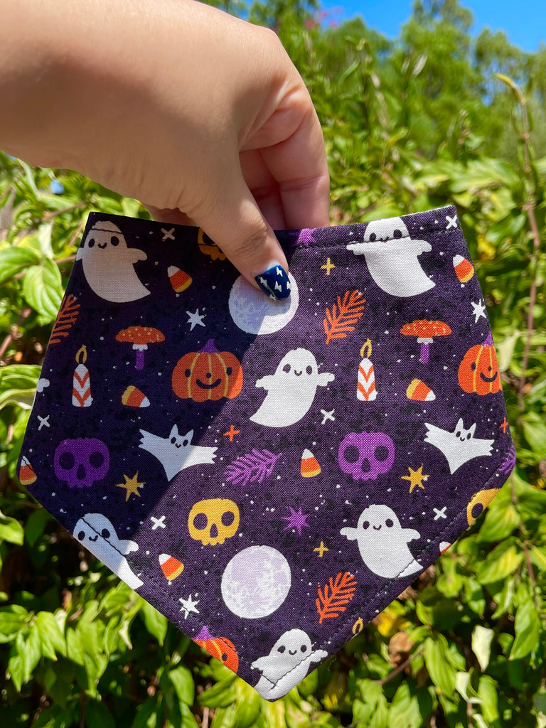 Hand holding festive Halloween bandana in front of leafy greenery; design includes mushrooms, bats, candy, and ghosts on a starry purple background.