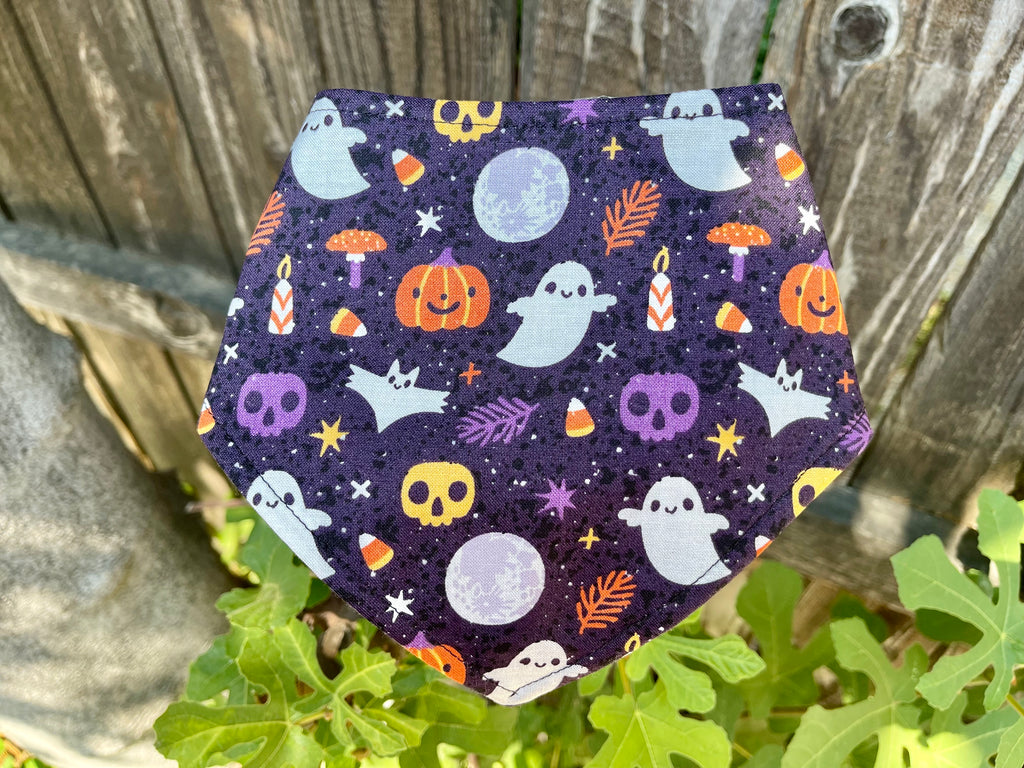 Halloween-themed dog bandana displayed on a wooden fence with green leaves; features ghost, pumpkin, candy corn, and moon print on a purple background