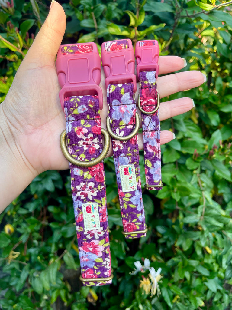 Hand holding small, medium, and large purple floral dog collars with pink and purple violet-like flowers, displayed in front of natural green ivy. Handmade, eco-friendly, organic cotton.