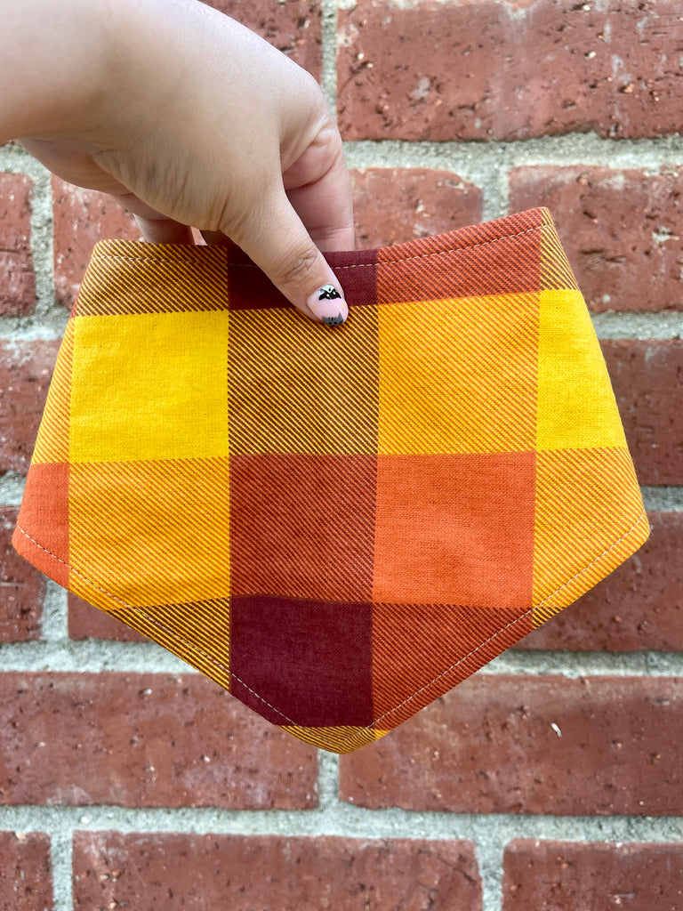Cozy fall bandana in yellow, orange, and maroon, adjustable with snaps held in front of a vintage brick background