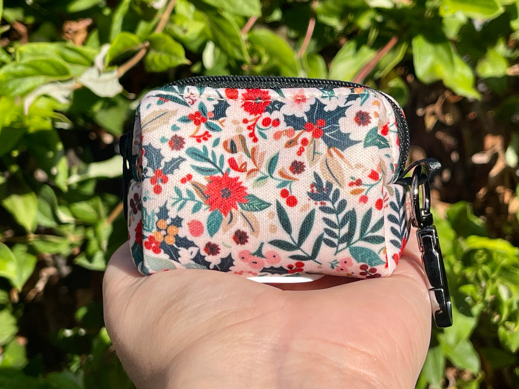 Holiday floral poop bag holder shown outdoors against leafy green plants, featuring red, cream, and green festive floral print