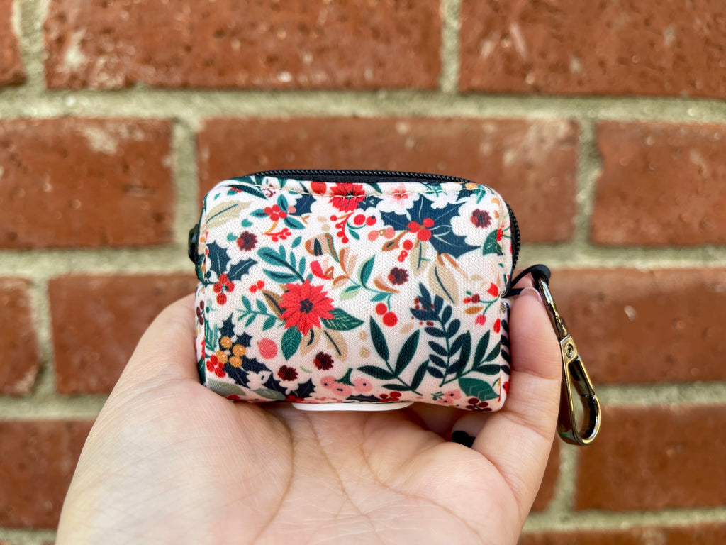 Hand holding a holiday floral poop bag holder with poinsettias, berries, and winter foliage on a cream background.