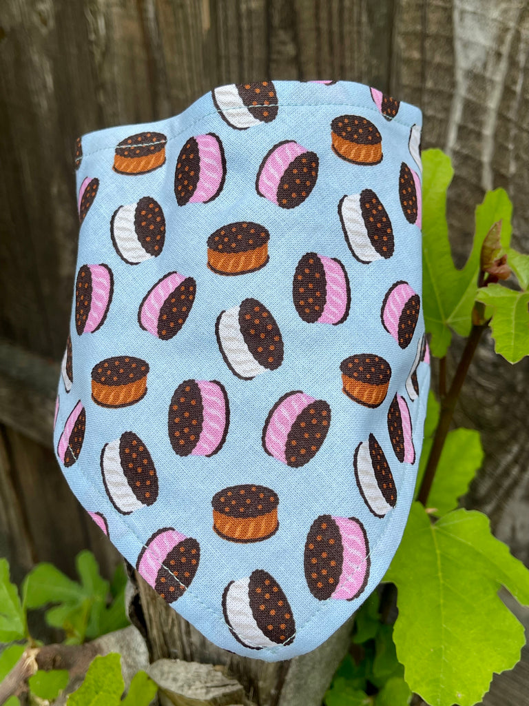 Light blue ice cream sandwich dog bandana displayed on a rustic wood post surrounded by fresh green leaves. Made from organic cotton.