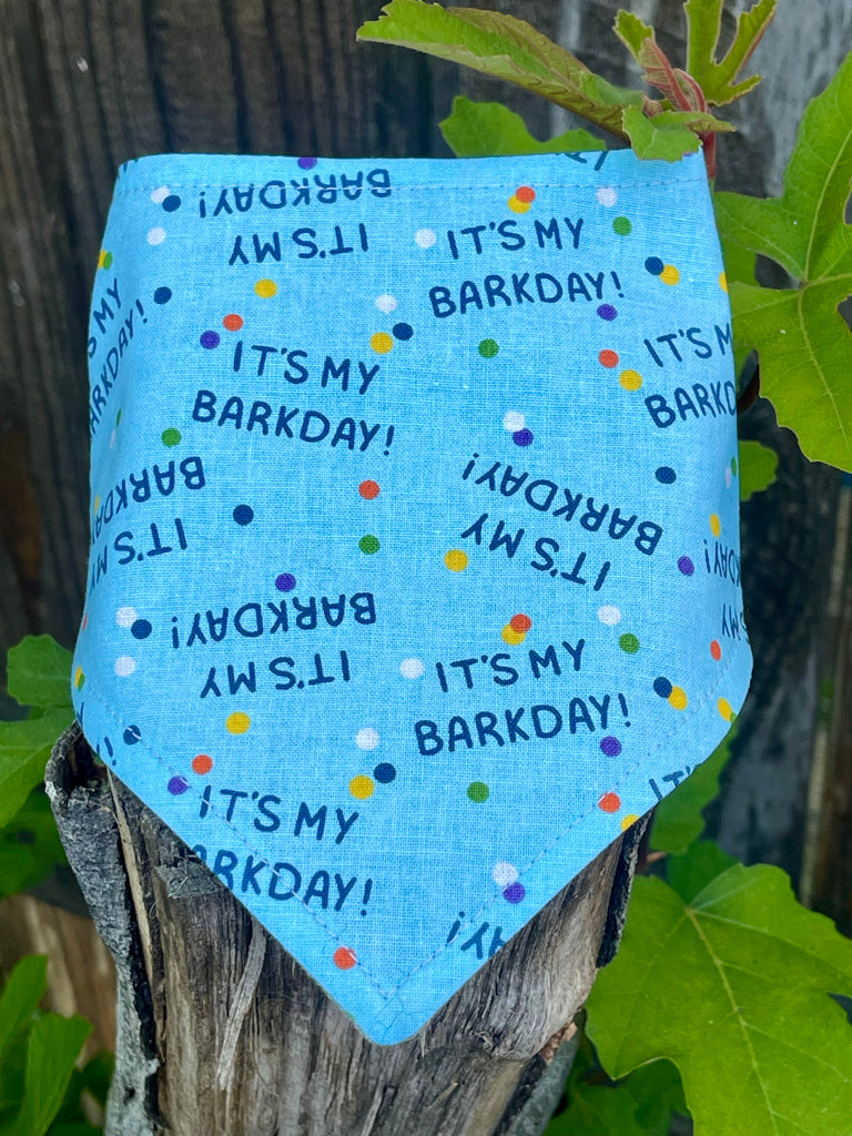 Close-up of handmade dog bandana with “It’s My Barkday!” design and confetti print. Styled on rustic tree stump with leaf detail.