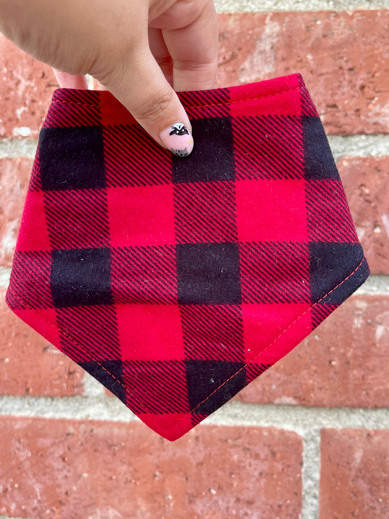 Hand holding a red and black plaid flannel dog bandana in front of a brick wall. Bold buffalo check pattern stands out