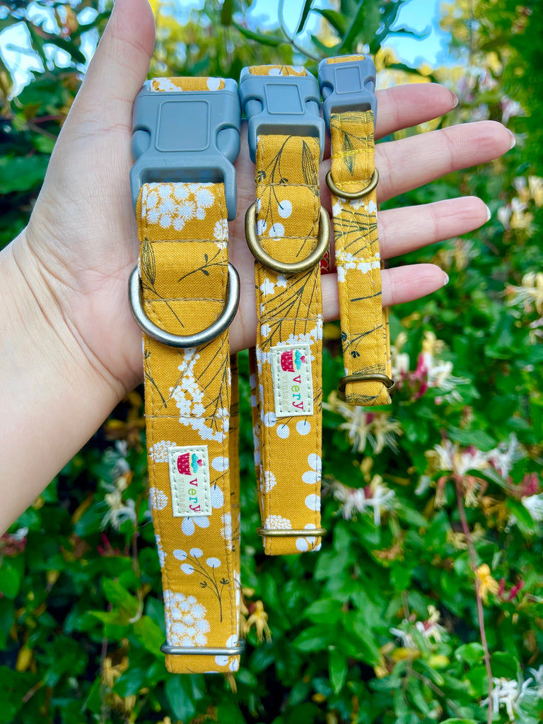 Handmade mustard yellow dog collar with vintage-inspired white and gray floral pattern, displayed outdoors in a lush garden setting. Adjustable and eco-friendly, made from organic cotton webbing with antique brass or antique silver hardware.
