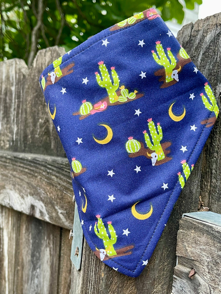 Western-style dog bandana in midnight blue with cacti and stars, hanging on a wooden fence for display.