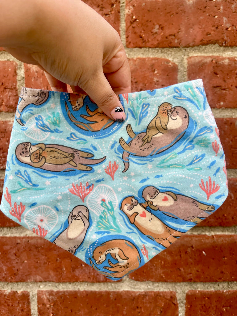 Hand holding a light blue dog bandana with otters floating paw in paw. Displayed against a brick wall backdrop