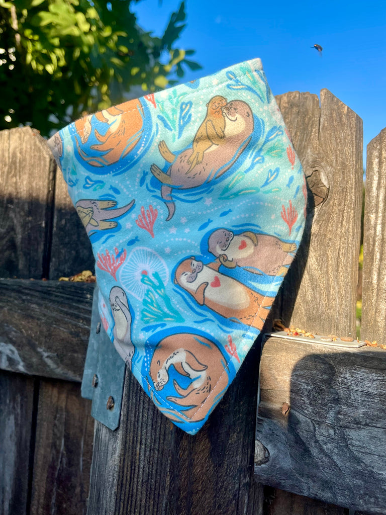 Playful dog bandana resting on a rustic wooden fence. Otter design visible with romantic light blue and soft cream backing