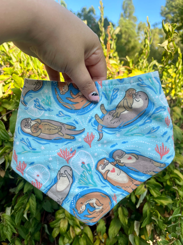 Otterly in Love bandana shown in front of leafy green plants. Features light blue fabric with otter couple and heart motifs