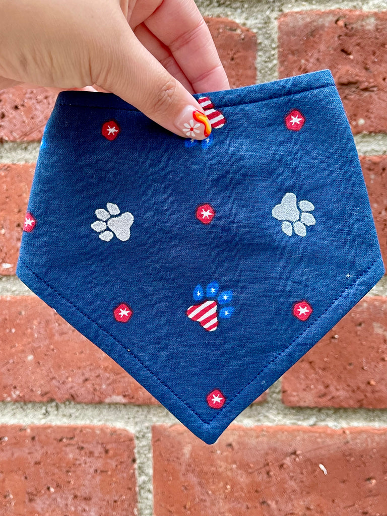 Navy dog bandana with scattered patriotic paw prints, styled outdoors with a vintage brick background for a fresh, festive product shot.