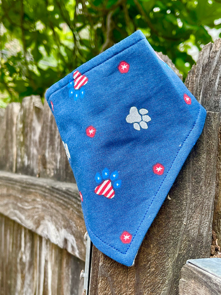 Navy blue dog bandana with red and white paw prints, draped neatly over a fence post, highlighting the fun patriotic print