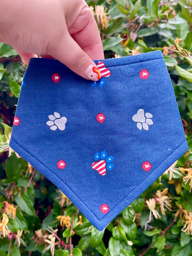 Hand holding a navy blue dog bandana with red and white paw prints, styled outdoors against a green leafy background, perfect for patriotic holidays.