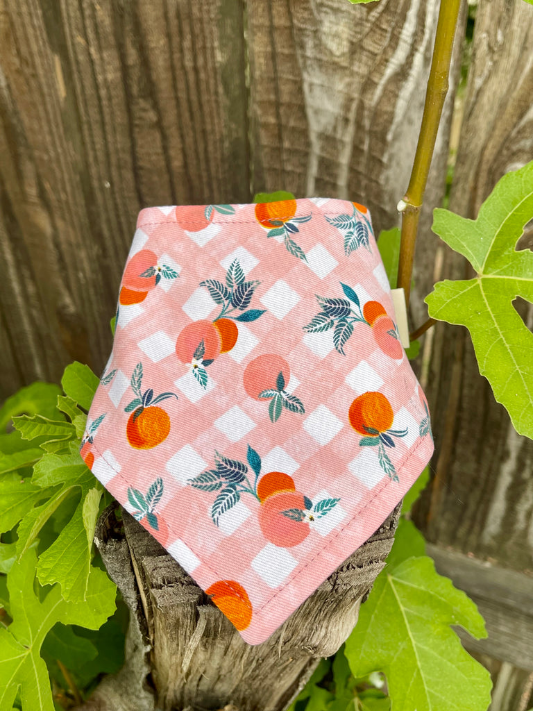 Plaid pink fabric with orange peaches and green leaves pattern on a wooden post with green leaves.