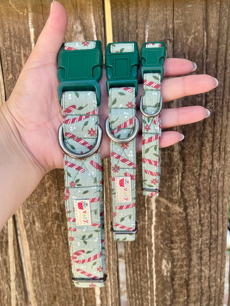 Three green with red candy canes organic cotton dog and cat collars held in hand, shown against a rustic wooden fence for texture contrast.