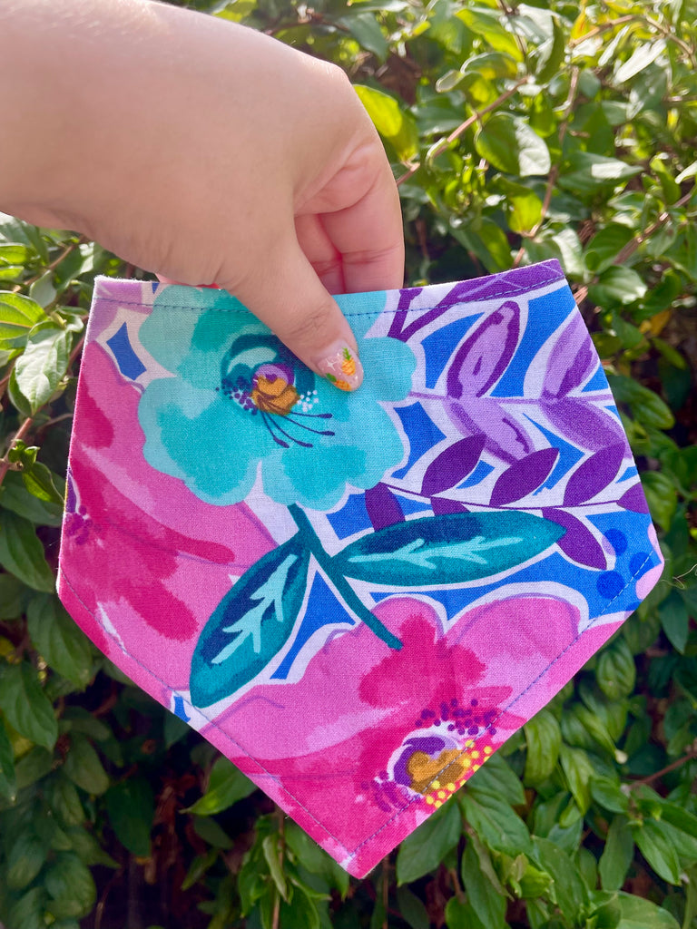 Floral print bandana held in hand against leafy hedge, featuring oversized aqua flower and bright pink and purple accents