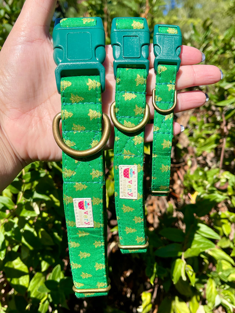 Hand holding three green Pine Trees collars in front of greenery – handmade Christmas dog collars with gold tree print and brass hardware