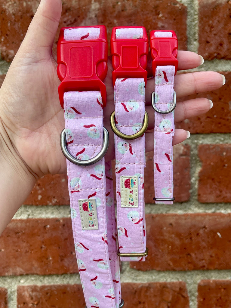 Hand holding pink Santa print dog collars with bright red buckles against rustic brick wall background
