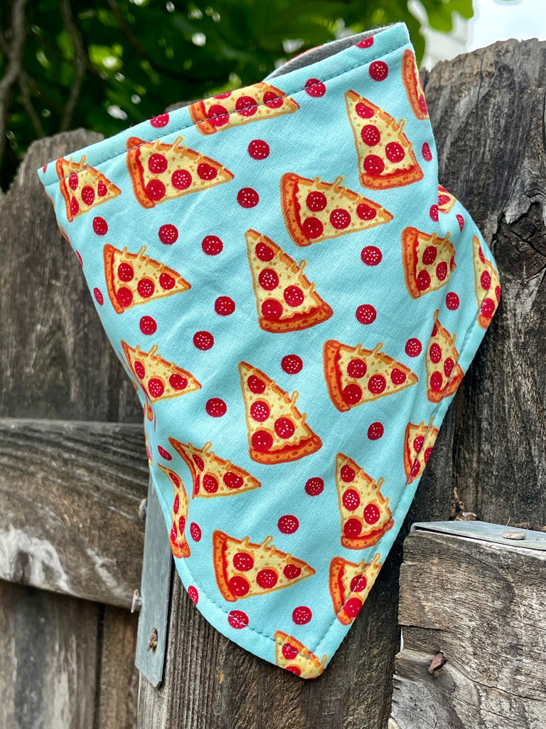Light blue pet bandana featuring pepperoni pizza slice pattern, displayed on rustic wooden fence, handmade eco-friendly dog accessory.