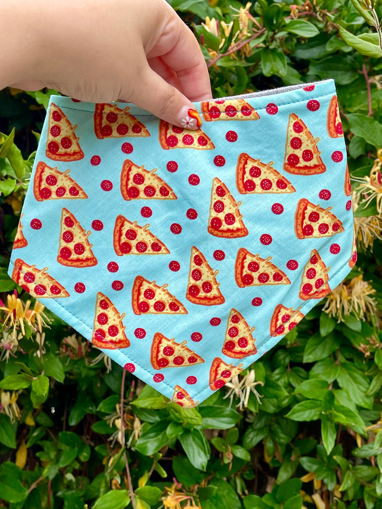 Close-up of blue pizza dog bandana held by hand, showing playful pepperoni slice design, eco-friendly handmade pet fashion. Handmade with love in Los Angeles, California.