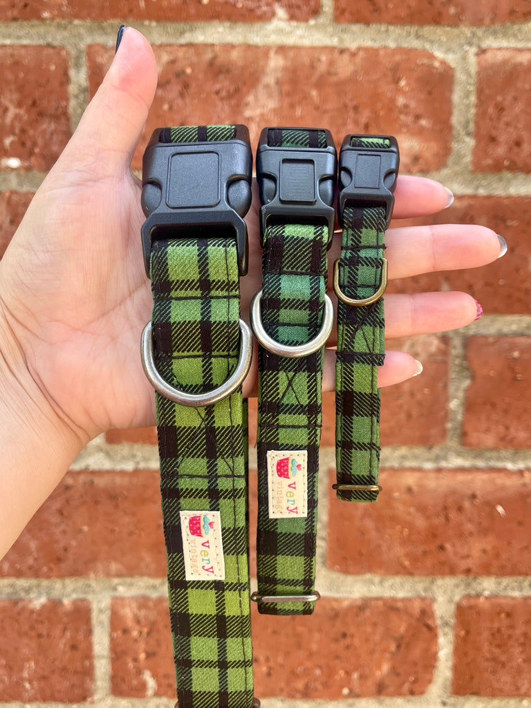 Green plaid dog collars in three sizes with black buckles, held in hand in front of red brick wall