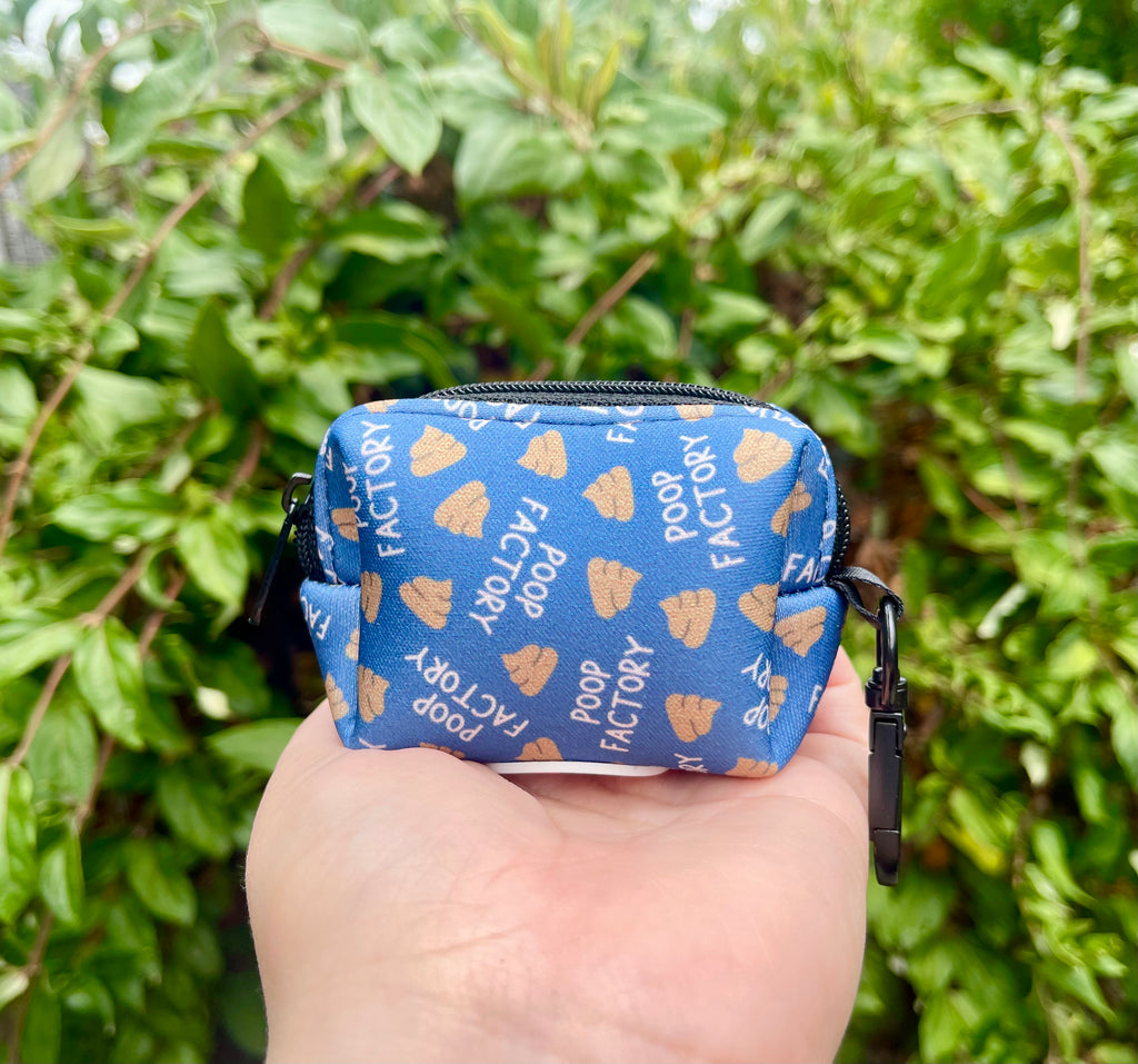 Blue poop bag holder with “Poop Factory” text, held in hand with green background