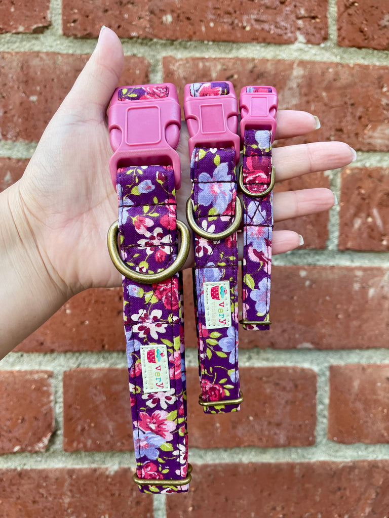 Three sizes of eco-friendly purple floral dog collars held by hand in front of a vintage brick wall. Features pink and purple flowers, perfect for spring. Handmade with organic cotton in Los Angeles. Choose from antique brass or antique silver hardware. Cat collars come with breakaway buckles.