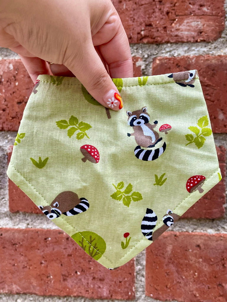 Handmade dog bandana with raccoons and mushrooms, displayed against a red brick background, emphasizing bright colors and fabric texture.