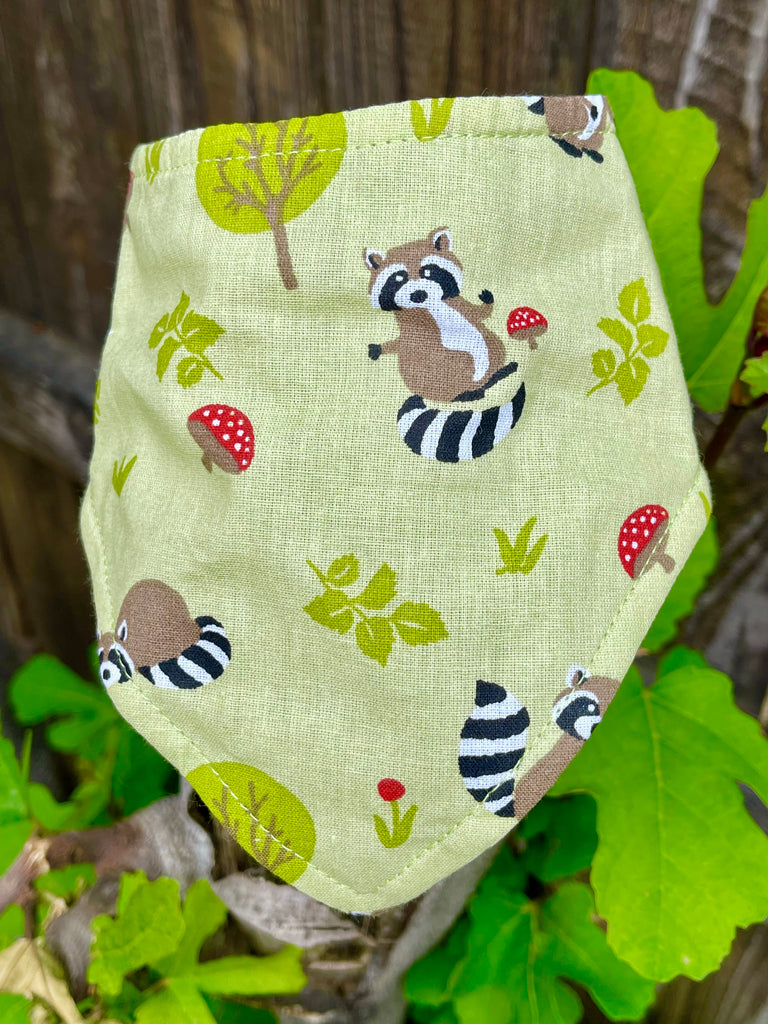 Close-up of raccoon and toadstool dog bandana propped on rustic wooden fence, highlighting the forest-inspired design details. Handmade with love in Los Angeles.