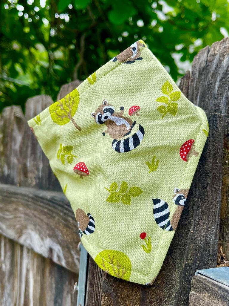 Light green handmade dog bandana featuring playful raccoons and red toadstools, styled draped on a wooden fence with greenery behind.