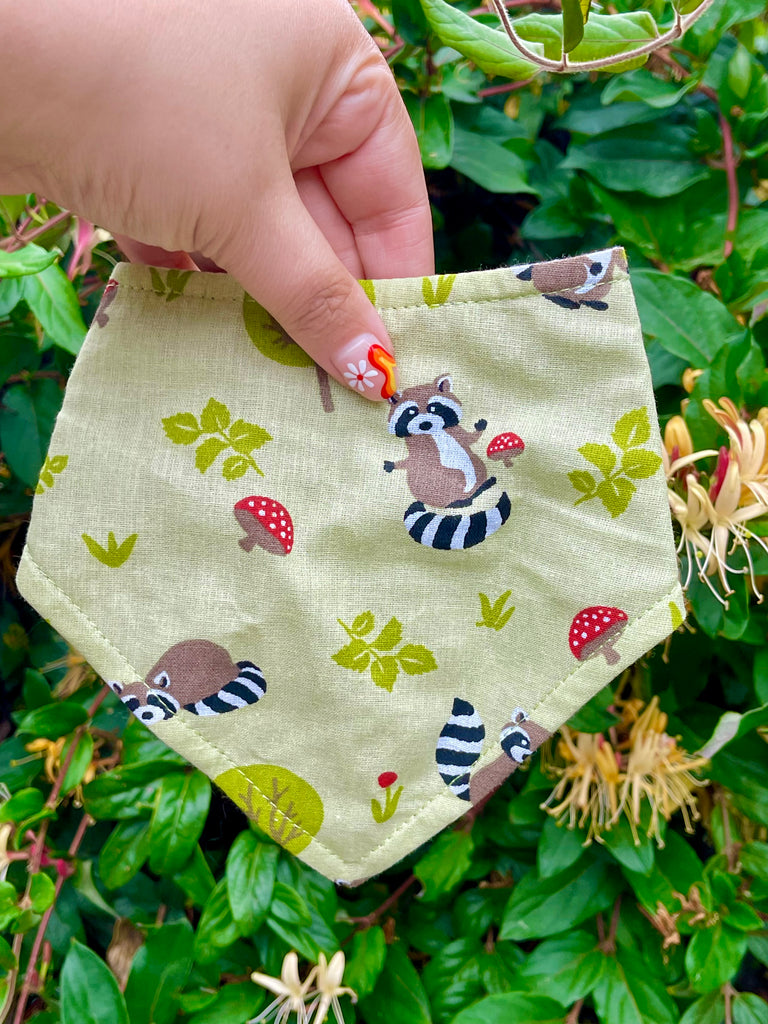 Light green raccoon and toadstool dog bandana held by hand against leafy garden background, showing playful woodland pattern up close.