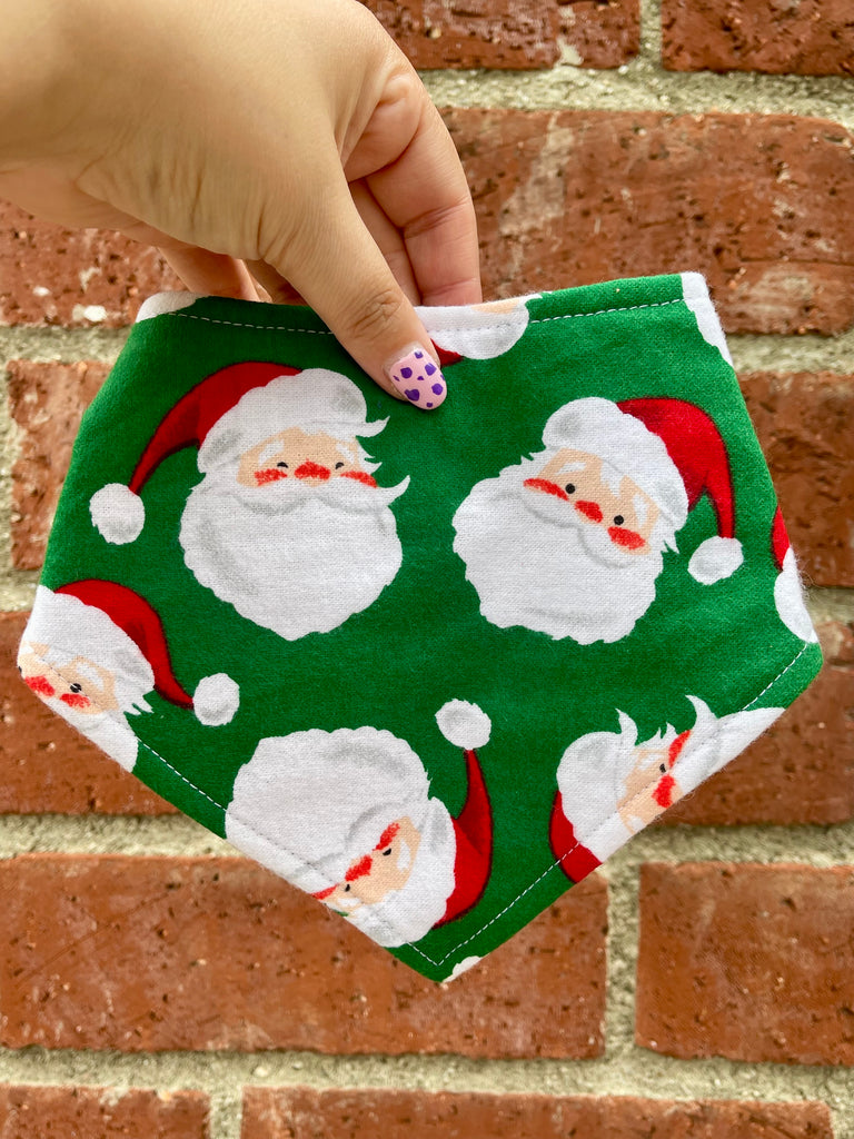 Close-up of Santa Paws holiday dog bandana, red and white Santa faces on green cotton, held in hand