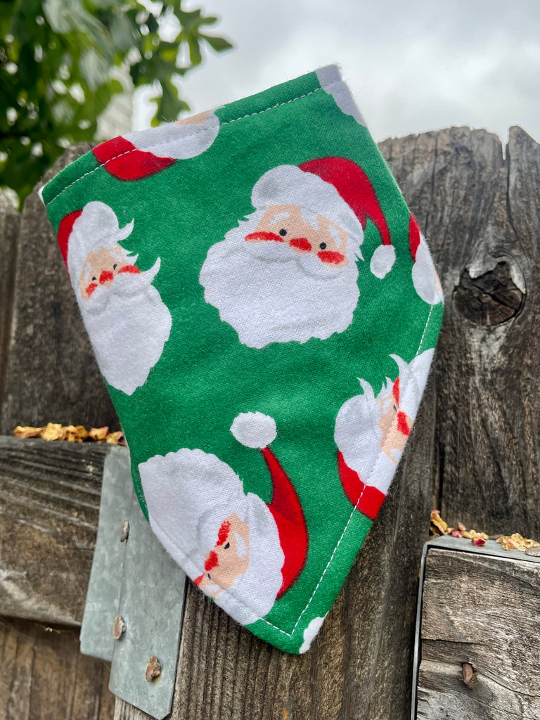 Dog bandana with Santa print on green fabric, displayed on wood fence, handmade holiday pet scarf