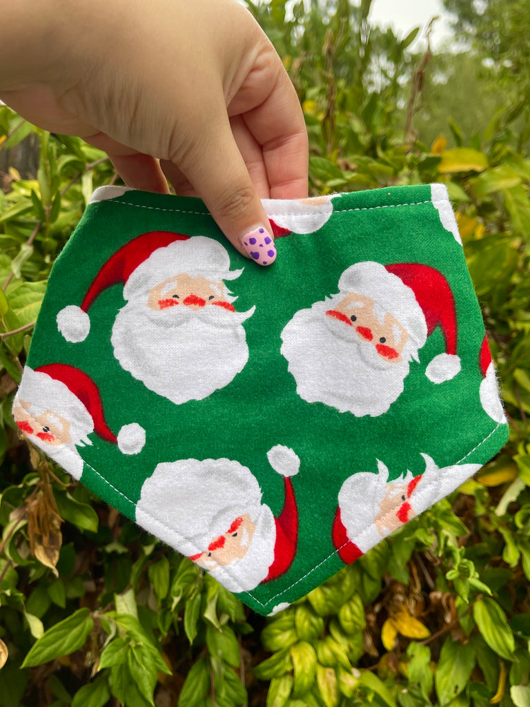 Green Santa dog bandana with jolly Santa faces, held outdoors with plants in the background