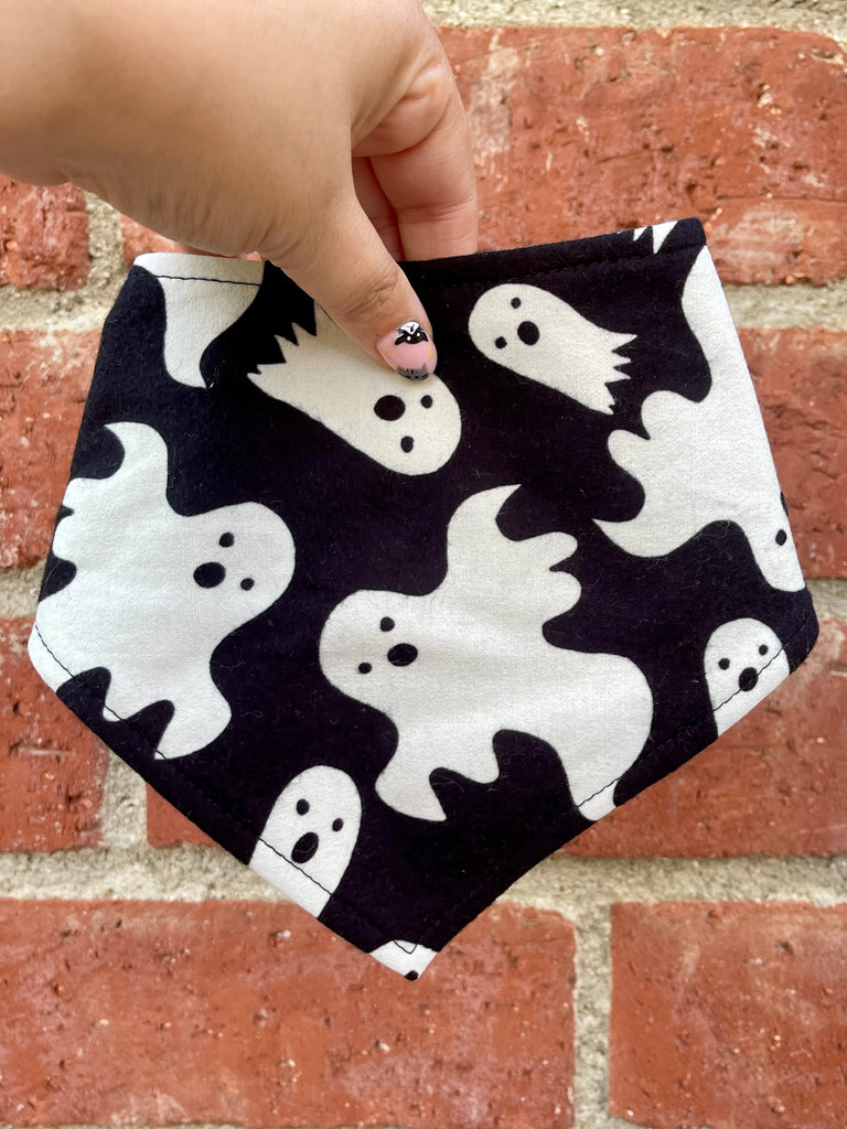 Hand holding a Halloween dog bandana featuring white ghost shapes on a black background. Brick wall behind adds warm contrast