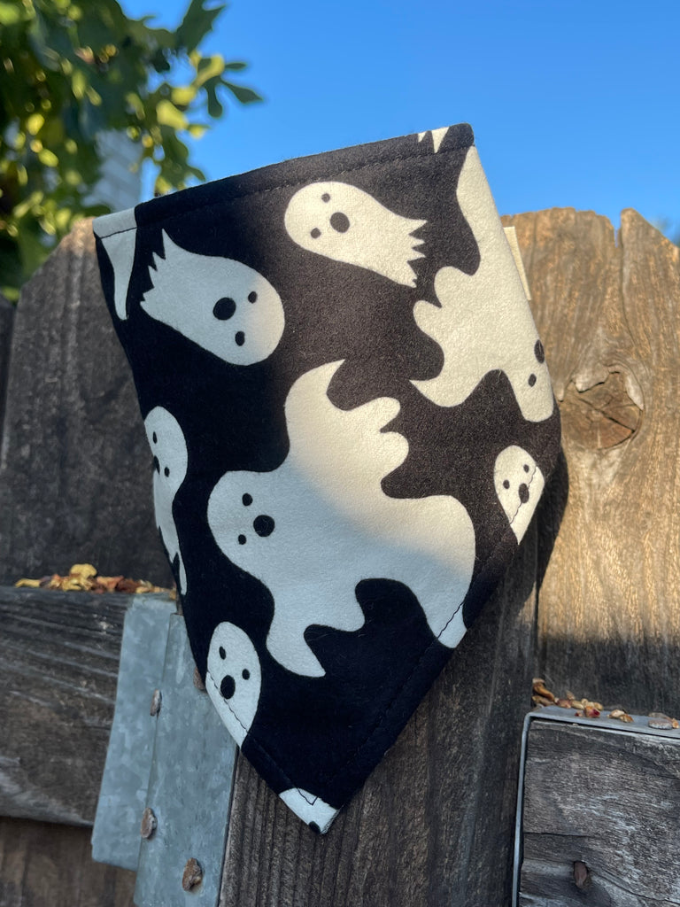 Spooky Ghosts bandana resting on a rustic wooden fence in soft morning light. Features playful white ghosts on black fabric