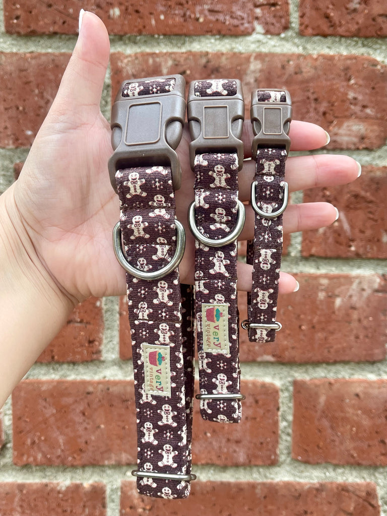 Hand holding three brown gingerbread dog collars with mocha buckles in front of red brick wall